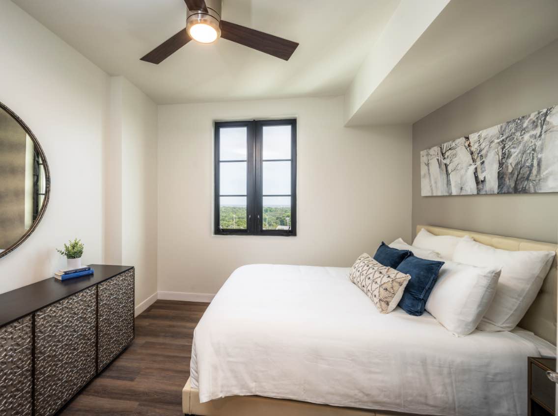 photo of bedroom with queen size bed with white comforter and pillows, window looking out to blue sky, large wall mirror and dark brown dresser